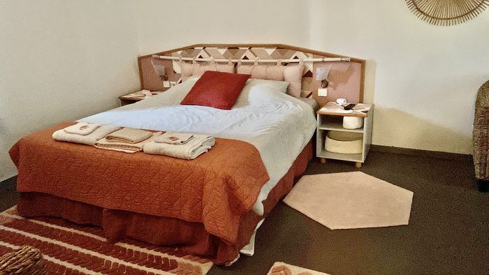 Chambre Bruyère : lit queen size, tête de lit en rotin, couvre-lit terre cuite, tapis tissé au sol.