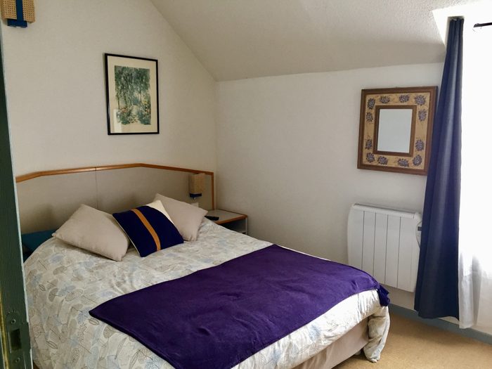 Chambre Campanule : lit double sous rampant, jeté violet, gravure encadrée et miroir au mur.