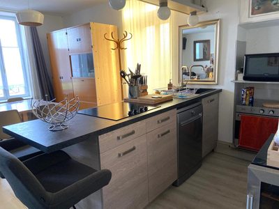Cuisine américaine : plan de travail sombre avec plaque à induction, lave-vaisselle, four et évier, prolongé par un bar et ses tabourets.