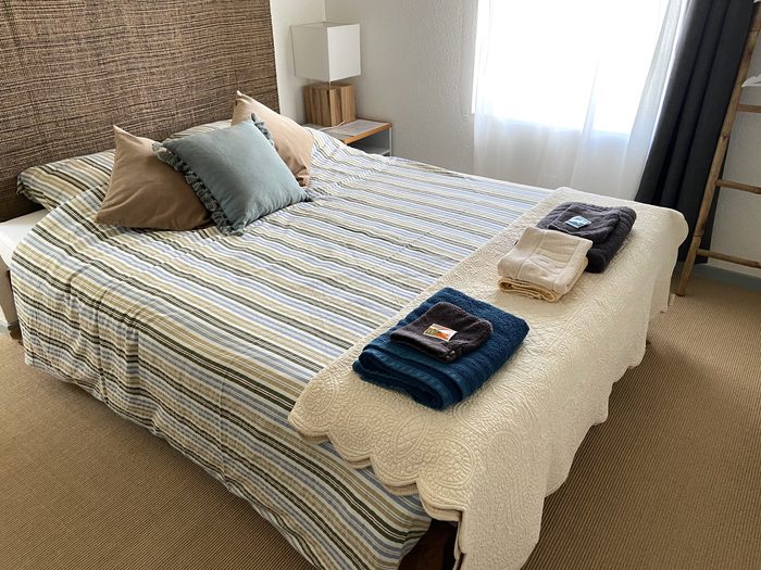 Suite Sapin : lit double habillé d'un couvre-lit rayé, tête de lit en fibre tressée, serviettes de toilette pliées au pied du lit.