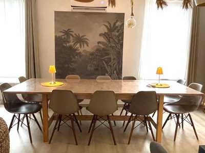 Longue table de bois entourée de douze chaises, deux petites lampes allumées dessus, devant une grande tenture botanique tendue au mur.